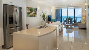 A modern kitchen with a large island, stainless steel appliances, and a dining area with a view of the ocean. - Vue Apartments Trinity Beach (Trinity Beach)