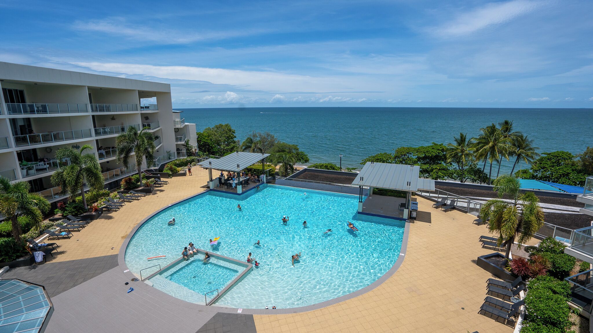 A hotel with a large pool surrounded by lounge chairs and palm trees, overlooking the ocean.
