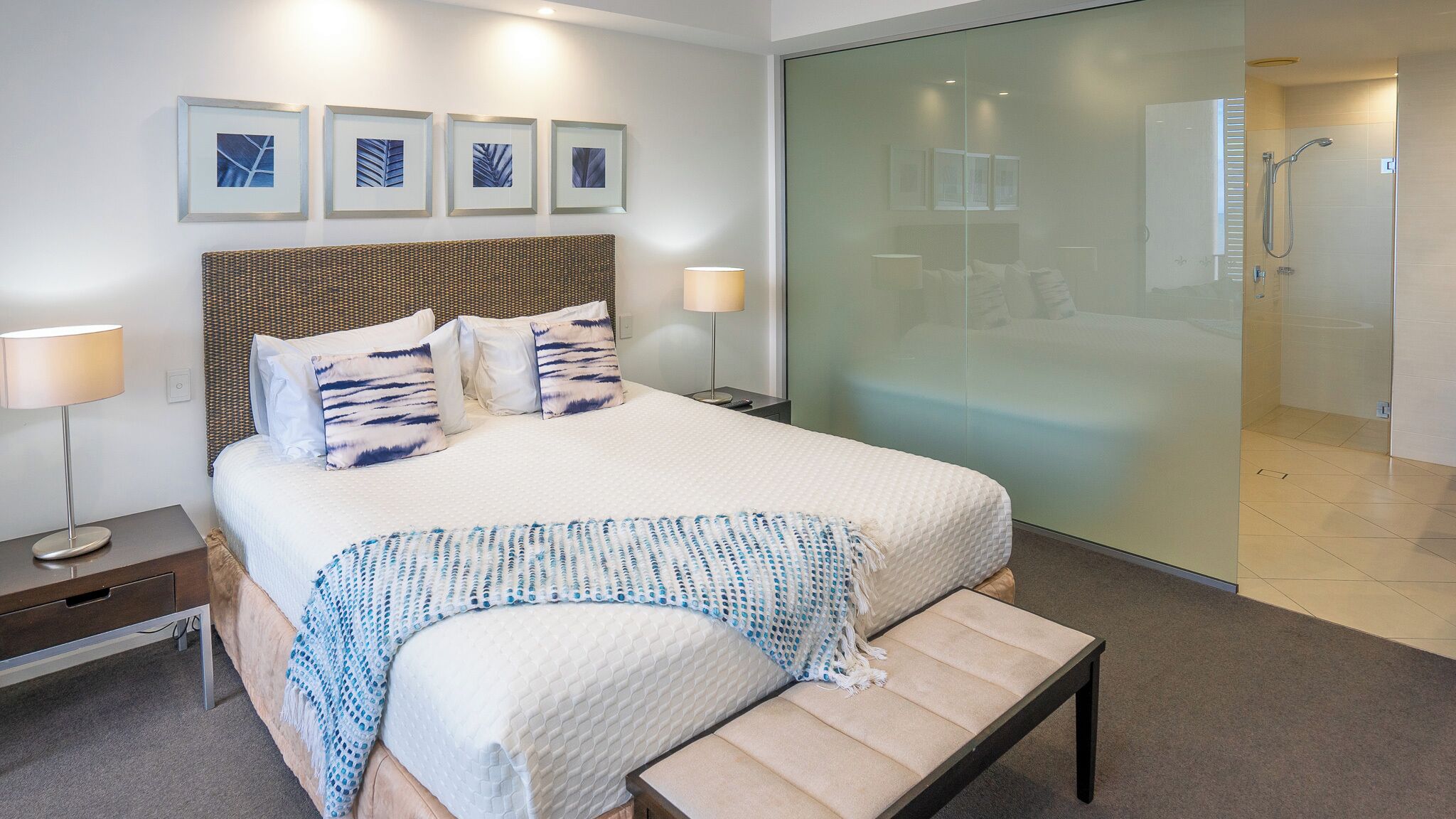 Ein modernes Hotelzimmer mit einem großen Bett, einem Nachttisch, einer Bank, einer Duschkabine aus Glas und gerahmten Kunstwerken an der Wand.