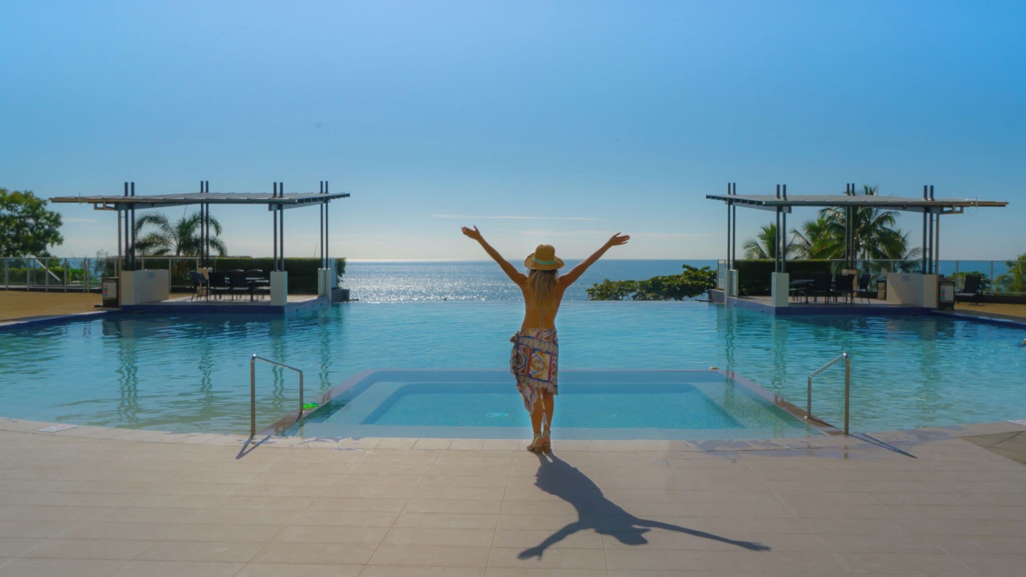 Una persona de pie al borde de una piscina, con los brazos levantados, mirando un cielo azul despejado y el océano.