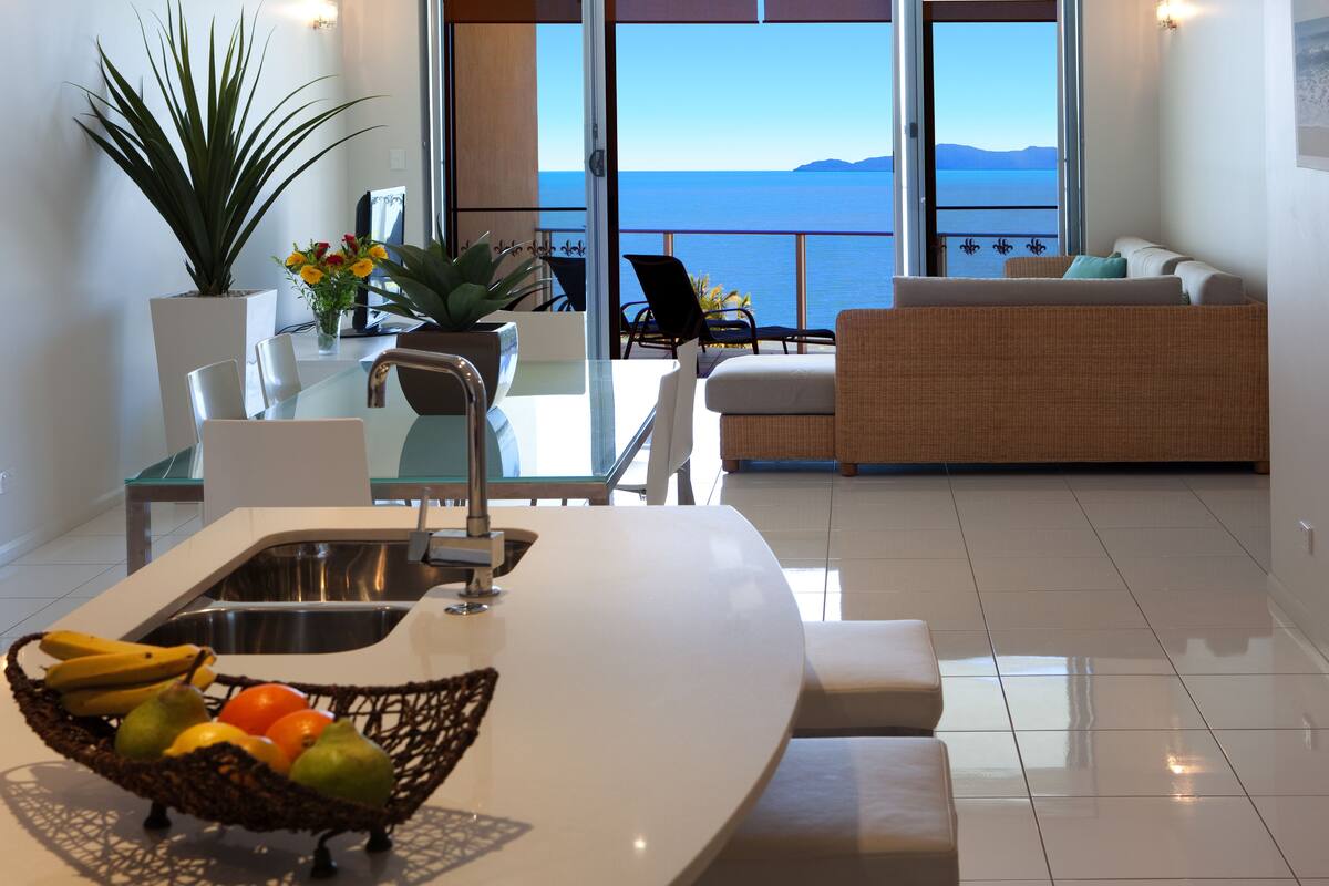 A modern kitchen with a large island, a dining area with chairs, a sofa, and a view of the sea.