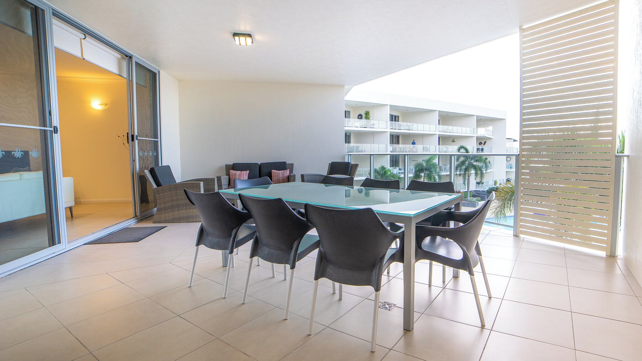 Un espace de salle à manger moderne avec une table en verre et des chaises noires, une porte-fenêtre coulissante donnant sur un balcon, et un intérieur blanc avec un luminaire au plafond.