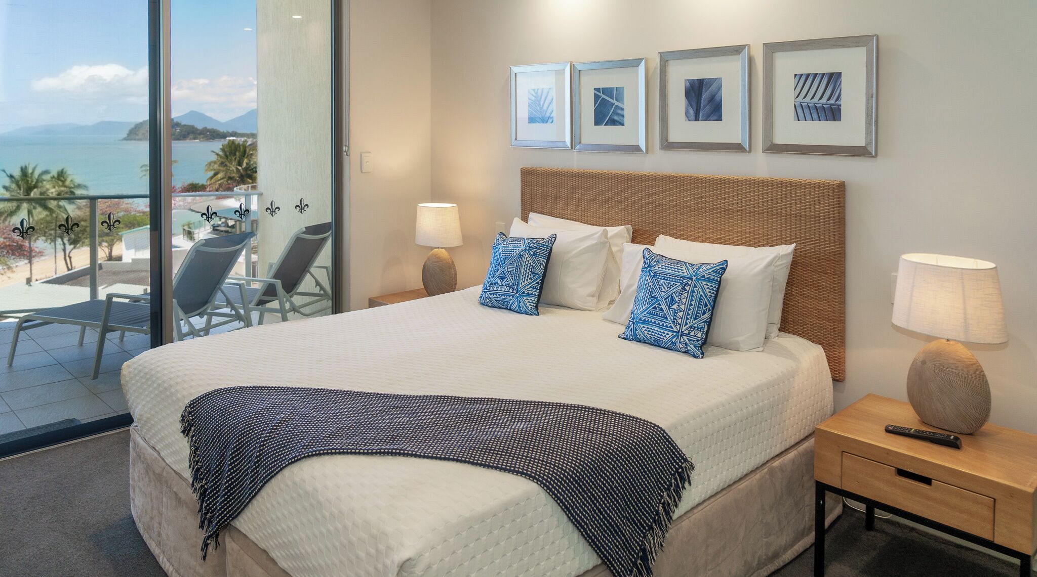 Ein Hotelzimmer mit einem großen Bett, zwei Kissen, einem Nachttisch mit Lampe, einer Fernbedienung und Blick auf den Ozean durch eine Schiebetür.
