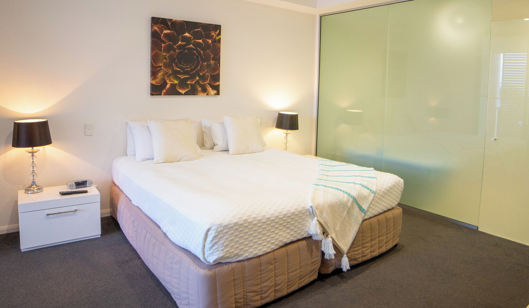 A neatly made bed with a white duvet, two bedside lamps, a remote control, a small bedside table, and a large abstract painting on the wall.