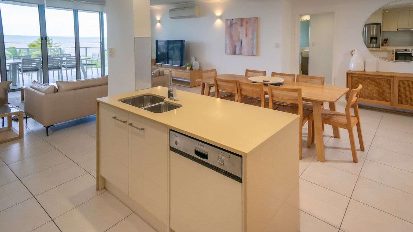 A modern kitchen with a dishwasher, a dining area with wooden chairs and a table, and a large window overlooking a balcony.