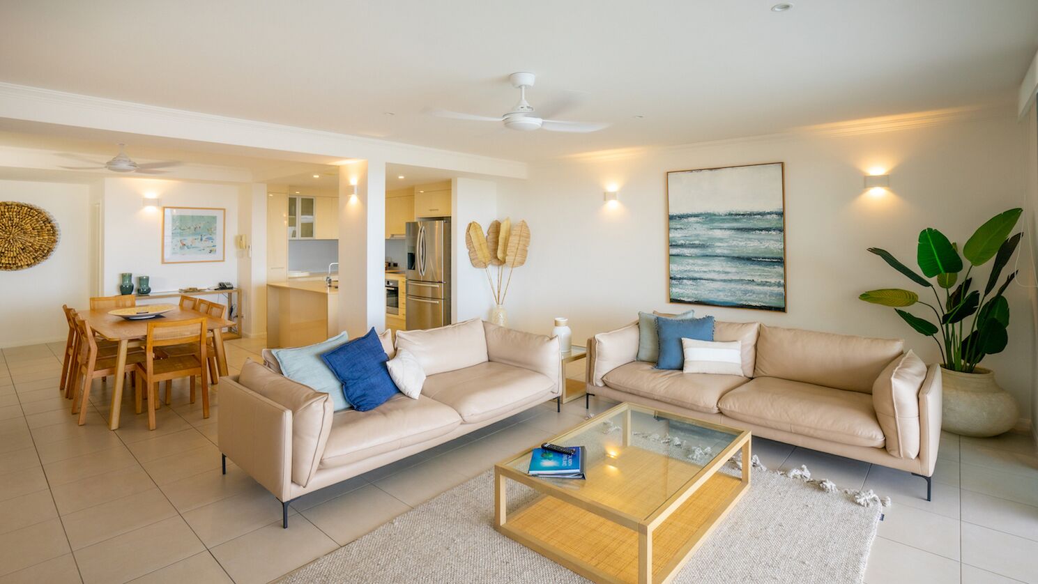 A spacious living room with two beige sofas, a glass coffee table, and a dining area with wooden furniture.