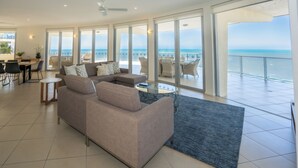A spacious living area with a large sofa, a coffee table, and a view of the ocean through large windows. - Vue Apartments Trinity Beach (Trinity Beach)