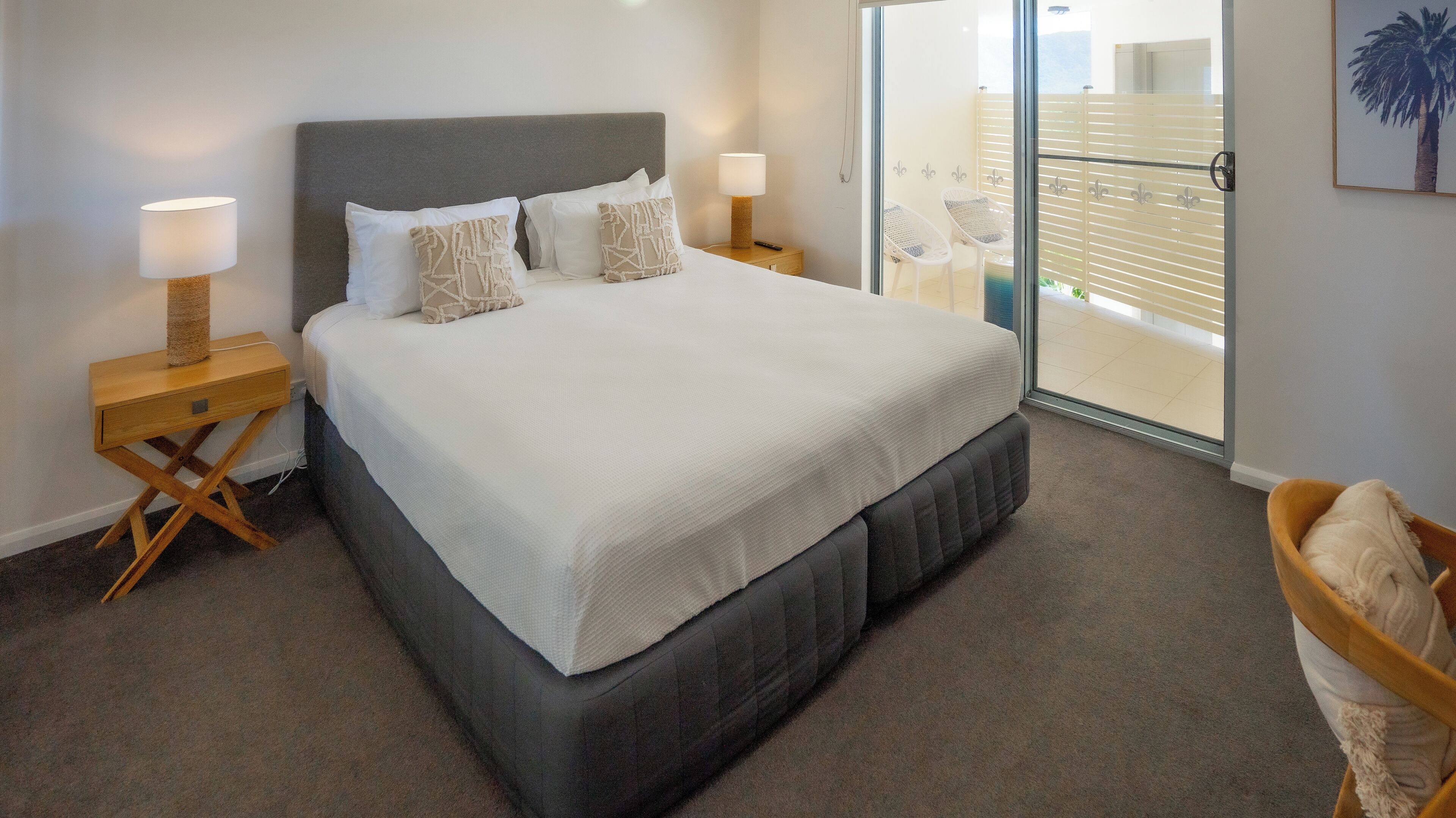 A neatly made bed with a grey headboard and white bedding, a bedside table with a lamp, and a view of the outside balcony through a glass door.