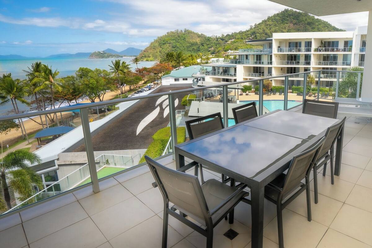 A balcony with a table and chairs overlooking a pool and beach.