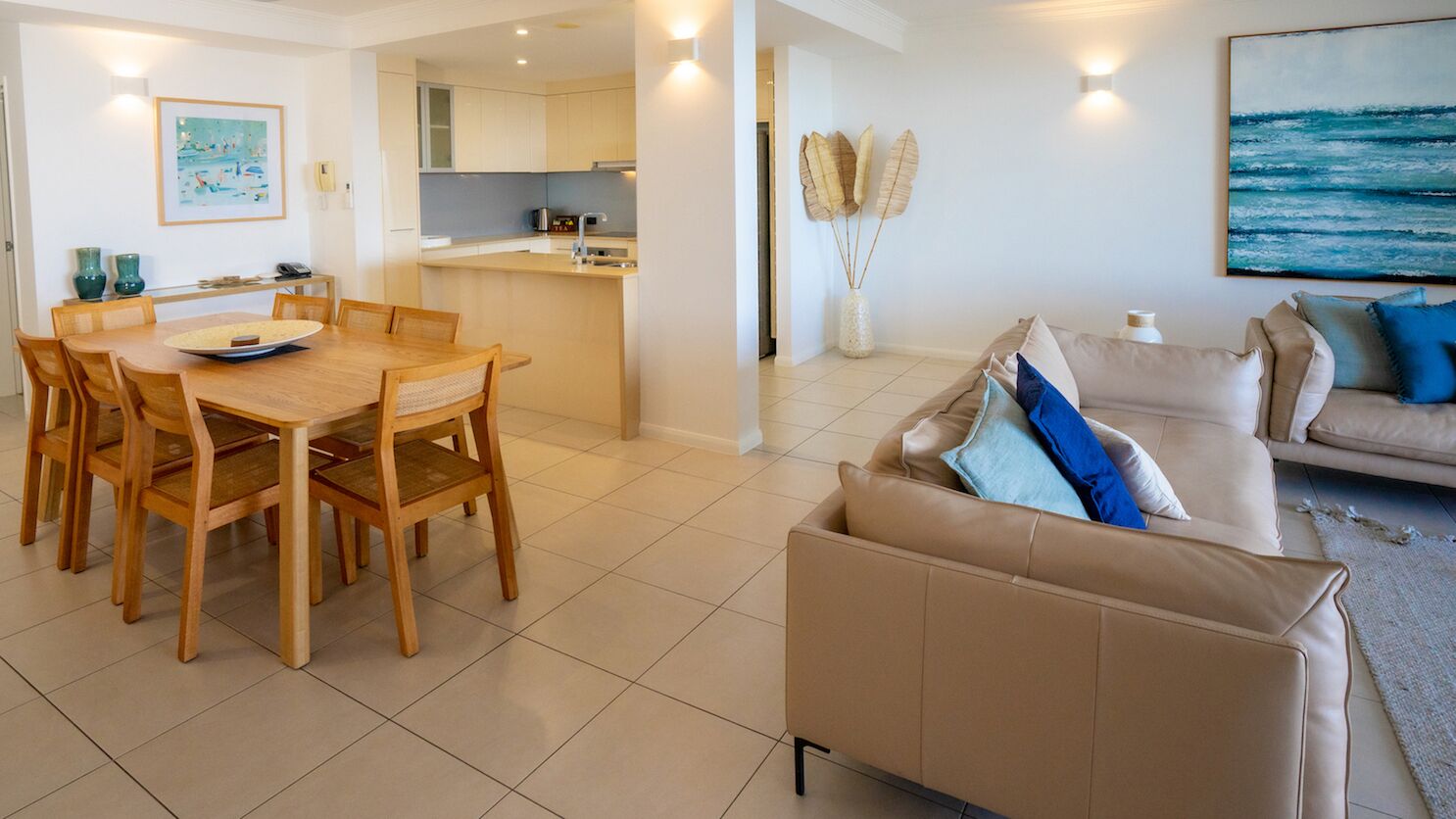 A modern living room with a beige sofa, a dining table with chairs, and a kitchen in the background.