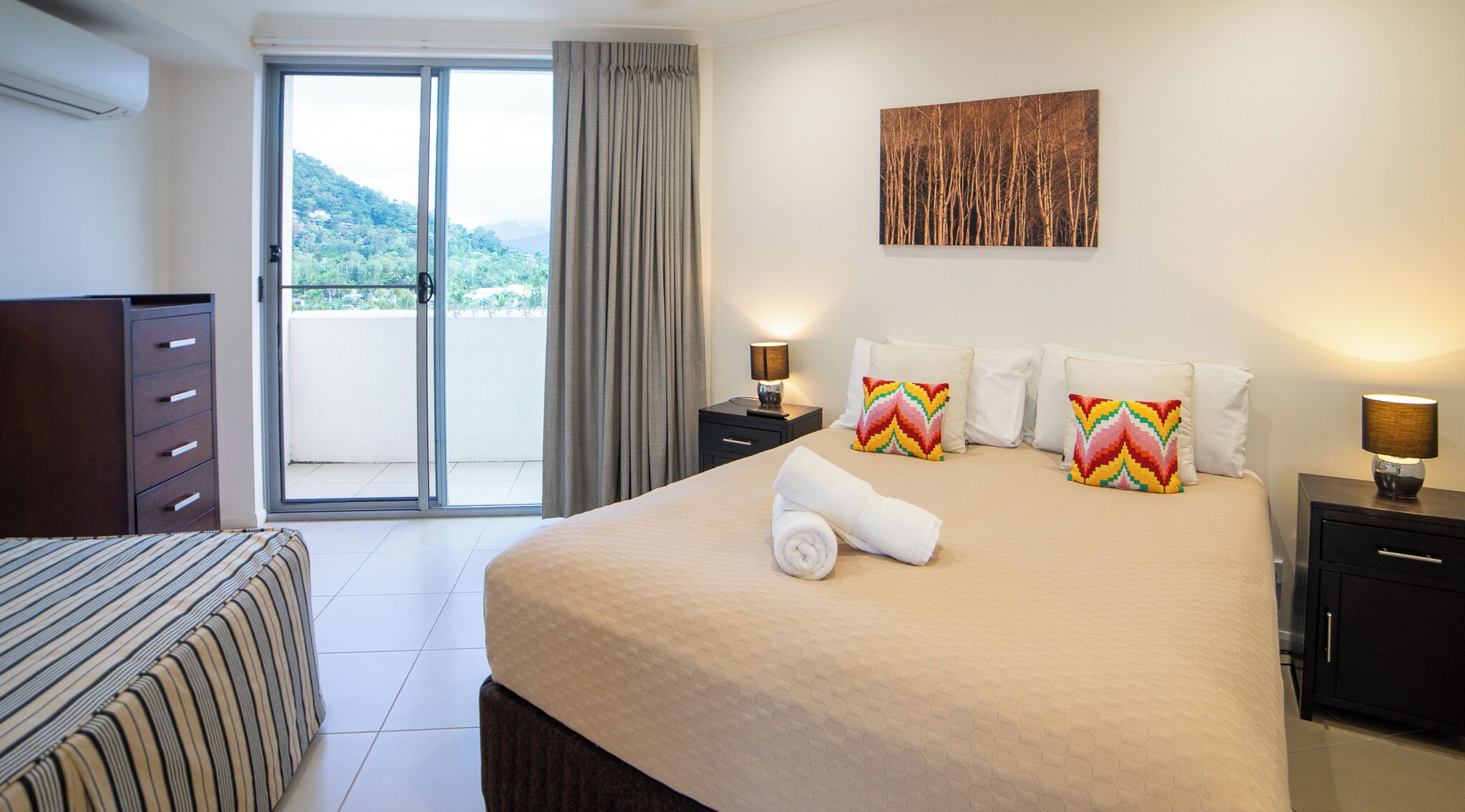 Une chambre d’hôtel avec un grand lit, une vue sur un balcon donnant sur un paysage montagneux, une commode en bois et une petite table avec une lampe.