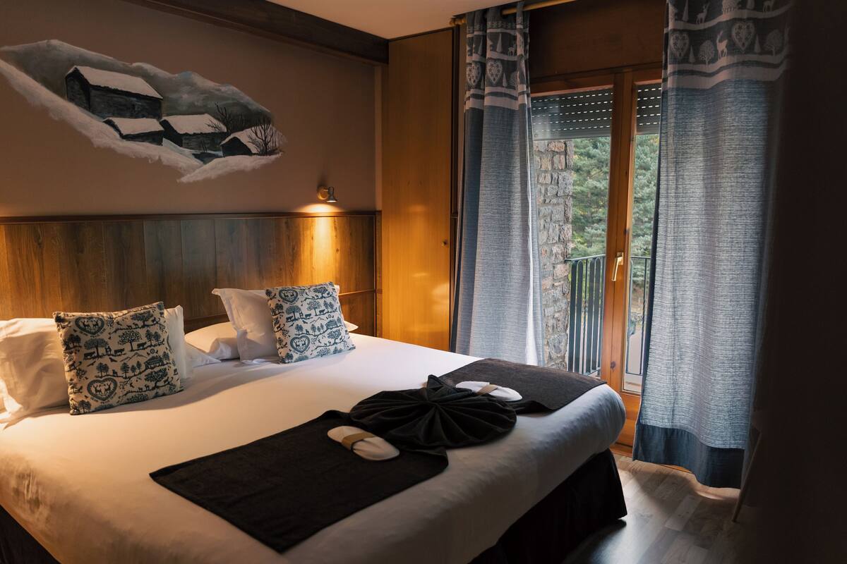 A hotel room with a large bed, wooden headboard, patterned pillows, a wall mural, and a balcony with curtains.
