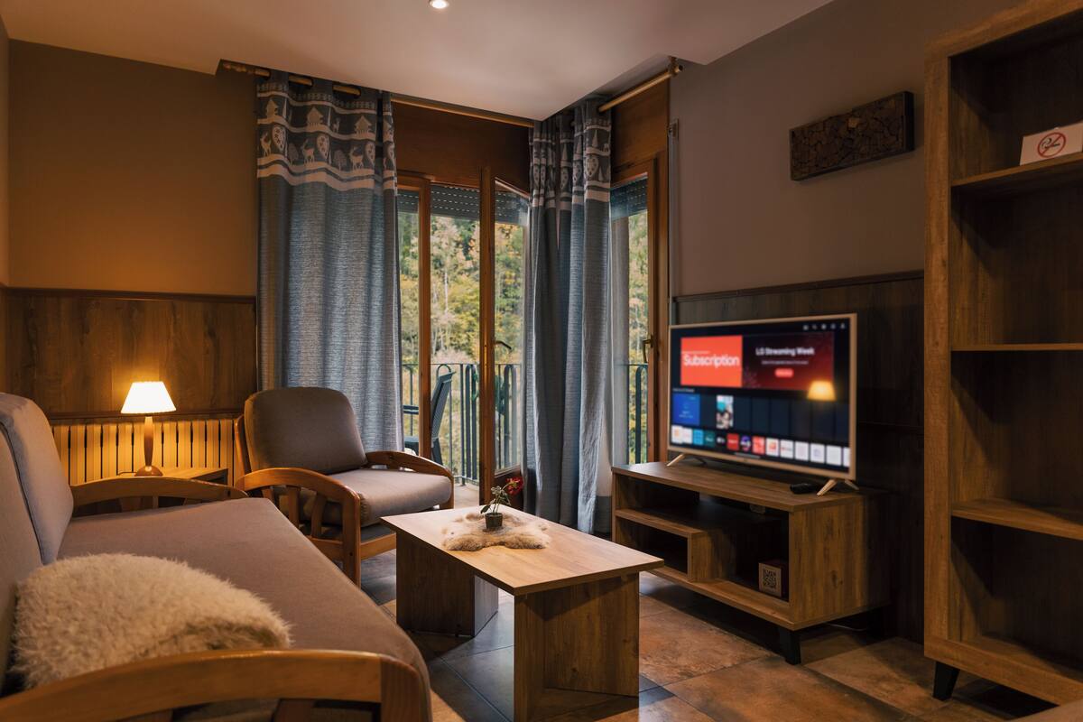 A cozy living room with a sofa, armchair, and coffee table. A television is mounted on a wooden stand.