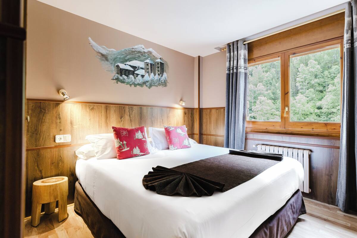 A hotel room with a large bed, wooden headboard, a painting of a snowy landscape, and a window with a view of trees.