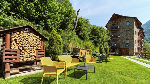 A rustic outdoor seating area with wooden furniture and a stack of firewood, set against a backdrop of lush greenery and a mountain. - Apartaments La Neu (Llorts)