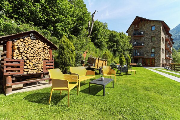 Un espace extérieur aménagé dans un style rustique, avec des meubles en bois et une pile de bois de chauffage, le tout mis en valeur par un décor de verdure luxuriante et une montagne en toile de fond.