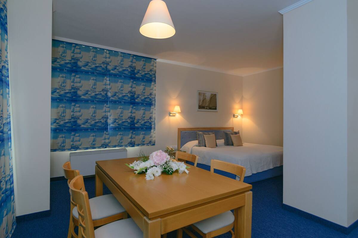 A hotel room with a wooden table, two chairs, a bed with pillows, a wall with a sailboat pattern, and two wall-mounted lamps.