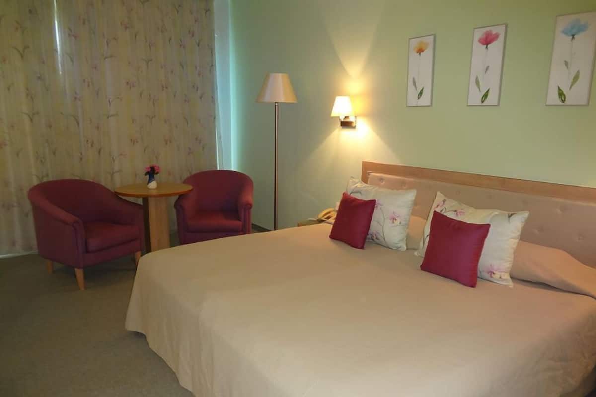 A hotel room with a large bed, two red armchairs, a small round table with a vase, and a lamp.