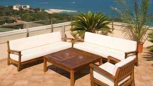 A terrace with a wooden table and two white sofas, overlooking a coastal view with the sea and distant buildings. - Boutique SPA Hotel Orchidea (Golden Sands)