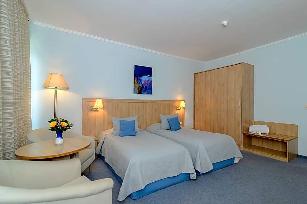A hotel room with two beds, a desk, a chair, a wardrobe, and a small table with a lamp and flowers.