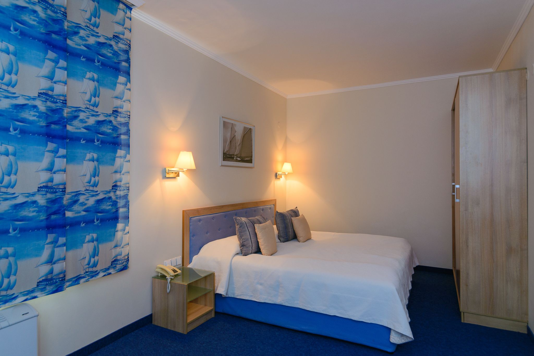 A bedroom with a bed, bedside lamps, a nightstand, a wooden wardrobe, and a wall with a sailboat pattern.