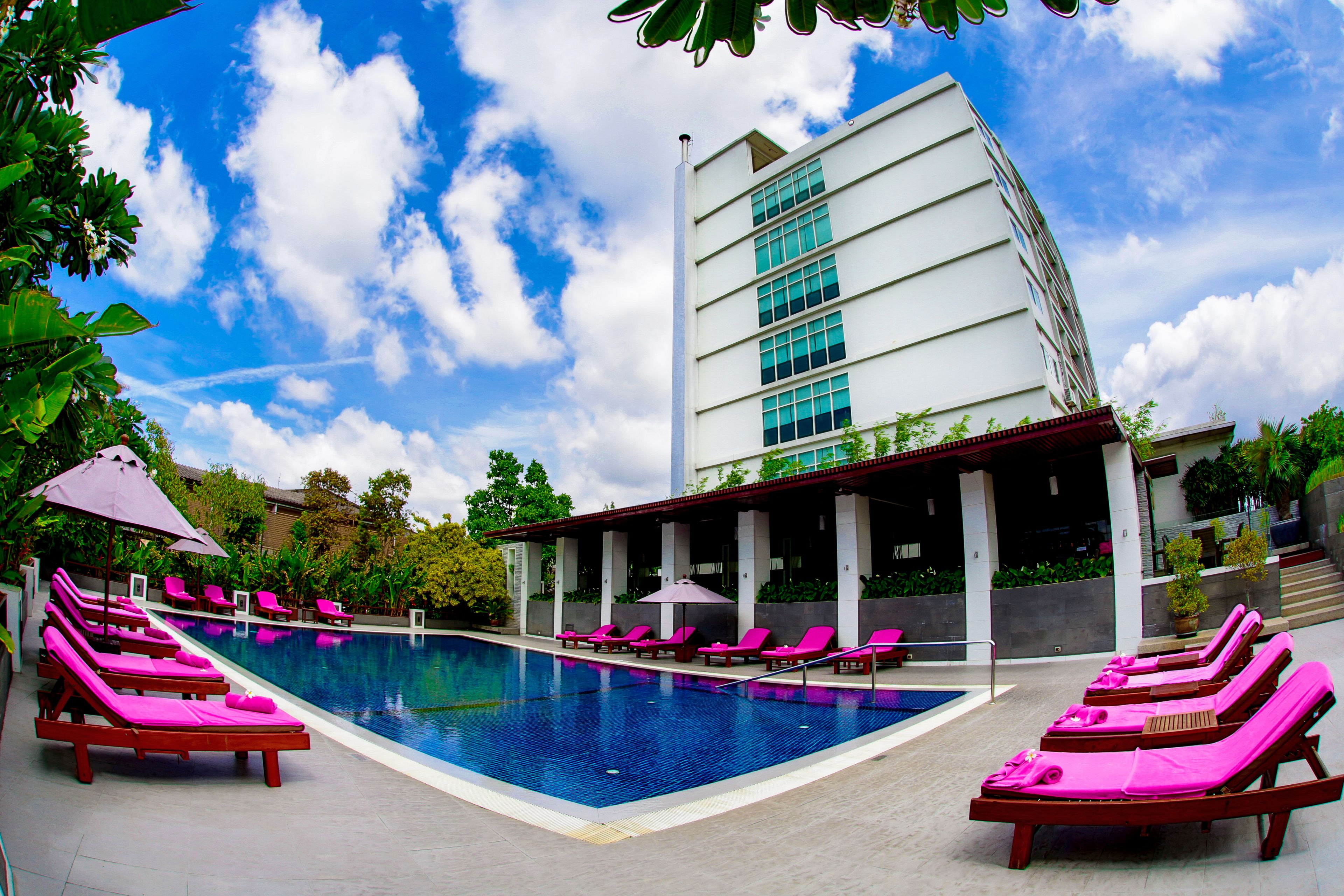 A swimming pool with pink lounge chairs and umbrellas, surrounded by greenery and a modern building.