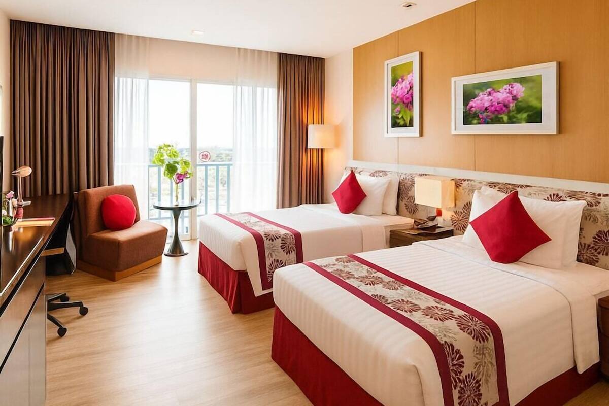 Ein Hotelzimmer mit zwei Betten, einem Schreibtisch, einem Stuhl, einem kleinen Tisch mit Pflanze und einem Balkon mit Vorhängen.