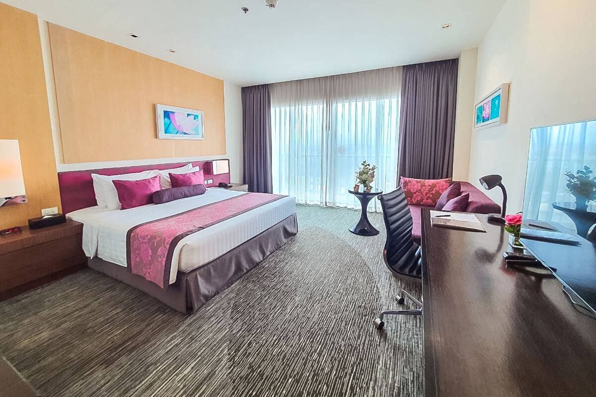 Ein Hotelzimmer mit einem großen Bett, einem Schreibtisch, einem Stuhl, einem kleinen Tisch mit Pflanze und einem fenster mit Vorhängen.