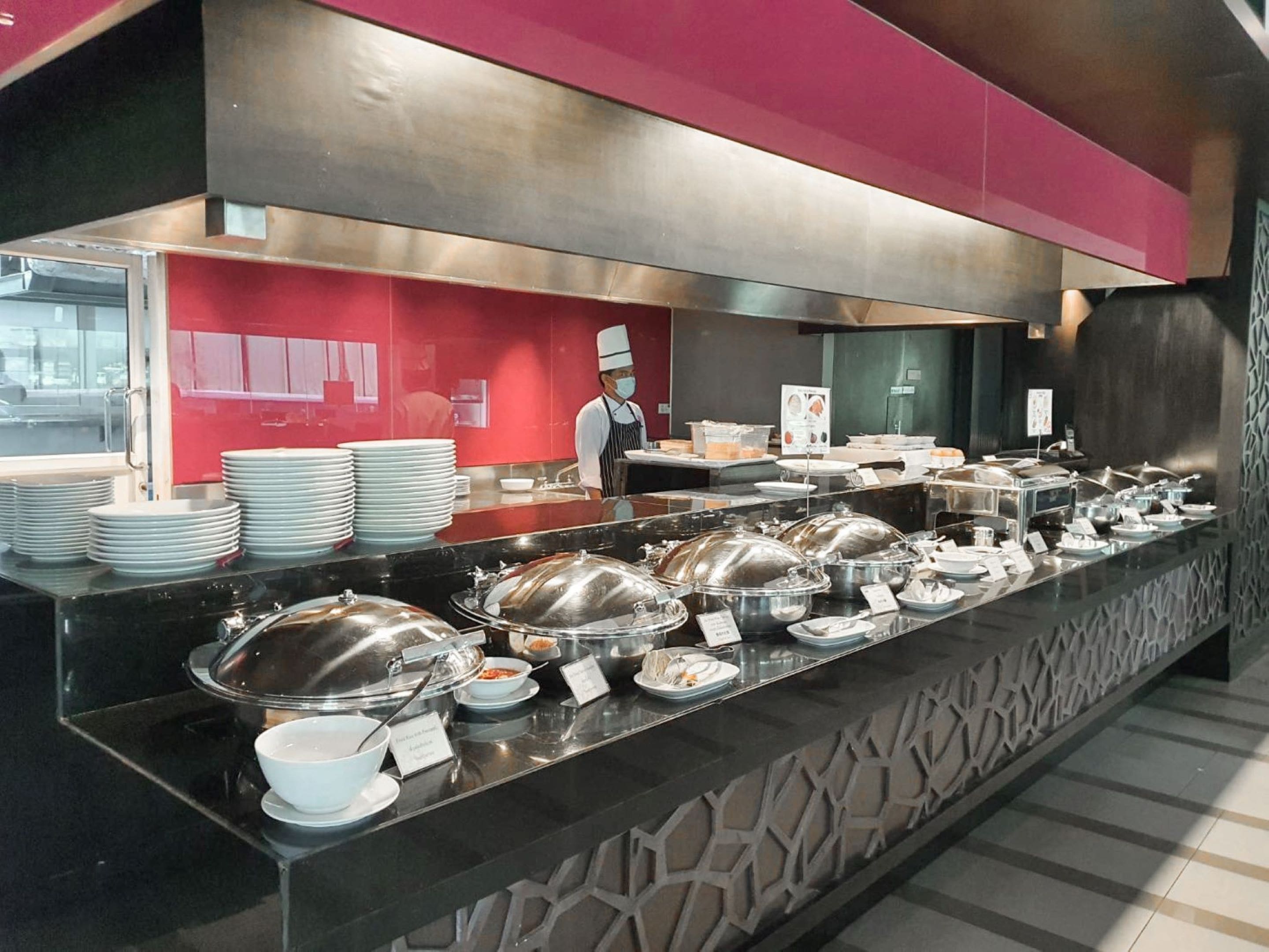 Un espace buffet avec plusieurs présentoirs de nourriture, un membre du personnel et un mur rouge agrémenté d’une fenêtre.