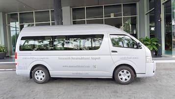 Una camioneta blanca estacionada frente a un edificio con grandes ventanales y un letrero que dice "Amarath Suvarnabhumi Airport".