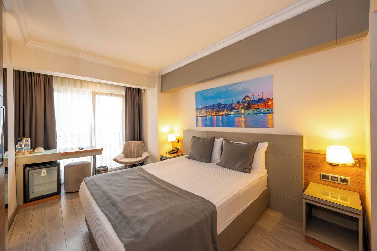 A hotel room with a bed, desk, chair, and a painting of a cityscape.