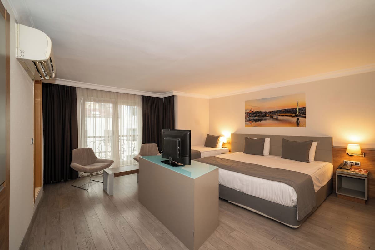 A hotel room with a large bed, a desk with a TV, a chair, and a view of a cityscape through the window.