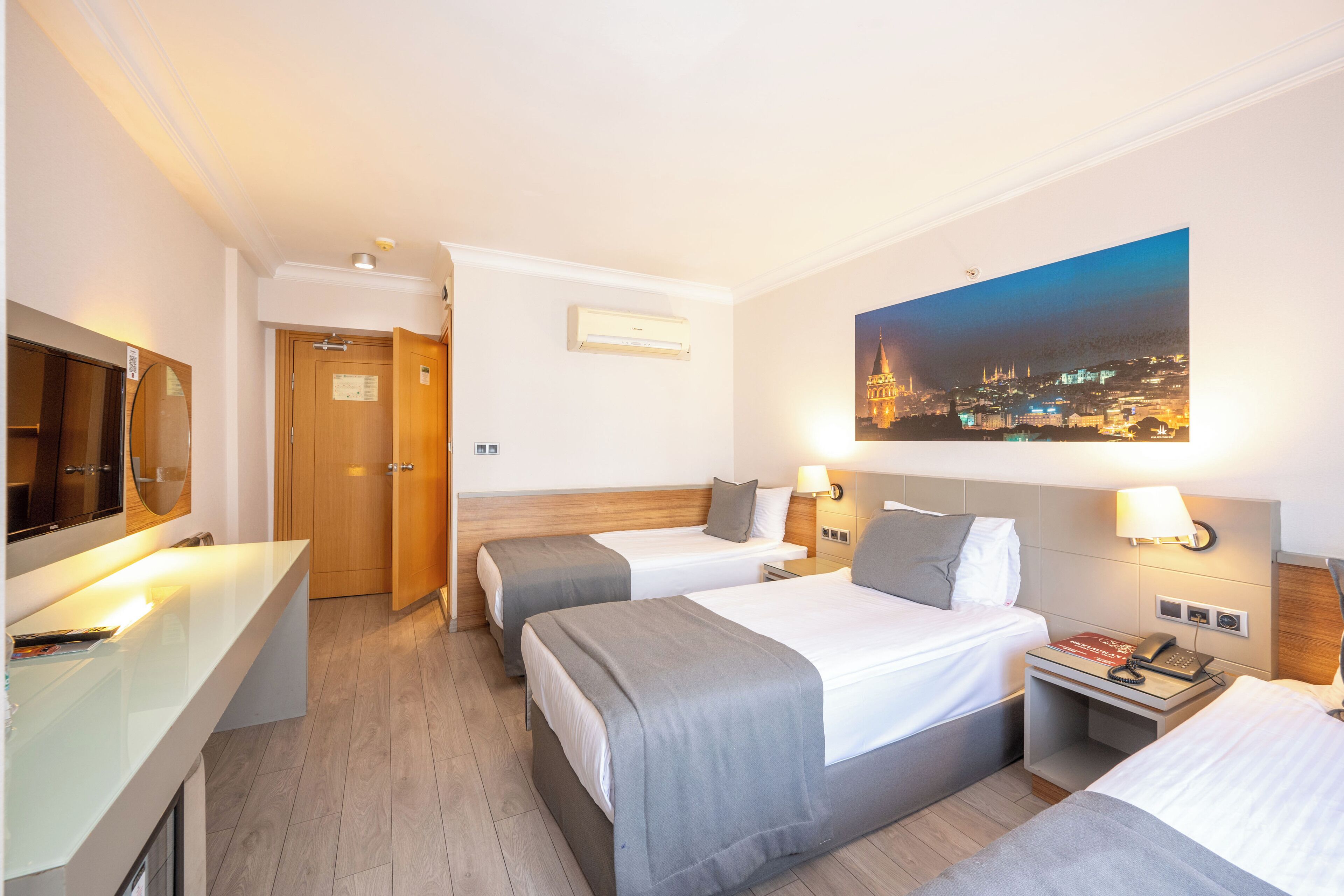 Een hotelkamer met twee bedden, een bureau met spiegel, een nachtkastje met telefoon en een stadslandschap op de muur.
