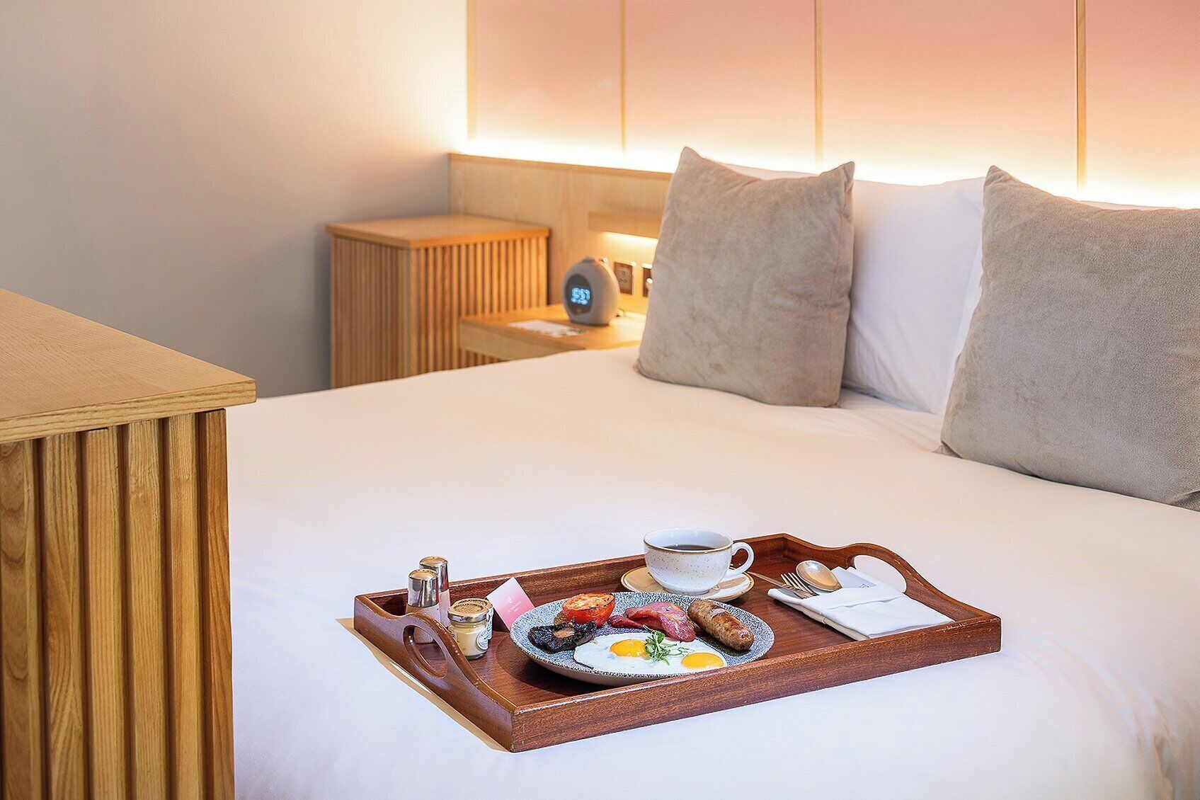 Ein ordentlich bezogenes Bett mit einem Tablett Frühstücksartikeln, darunter Eier, Speck und Toast, in einem Hotelzimmer mit einem Holzkopfteil und einem Nachttisch mit einer Lampe.