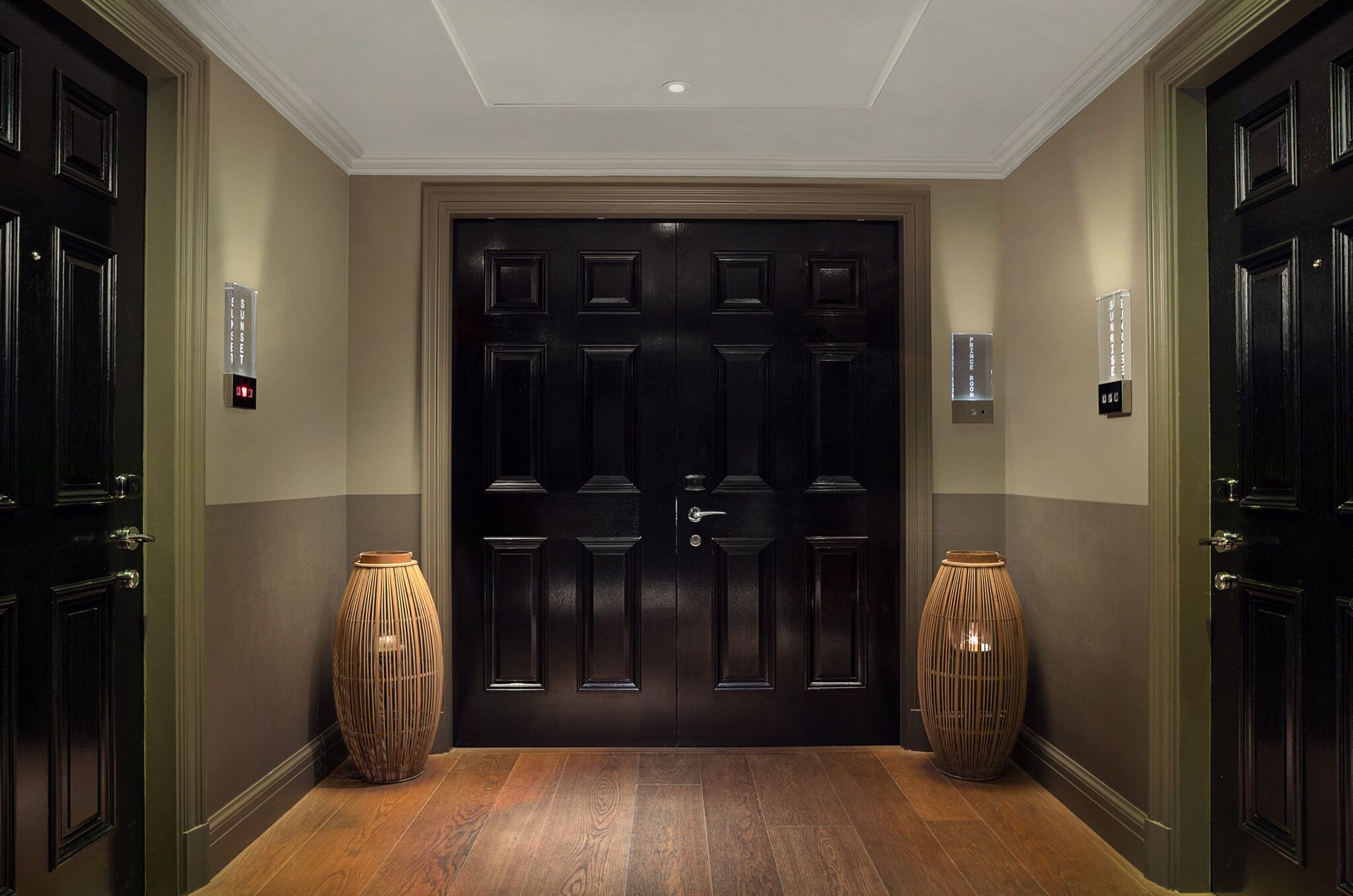 A hallway with a dark wooden double door, two wall-mounted light switches, and two large vases on either side.
