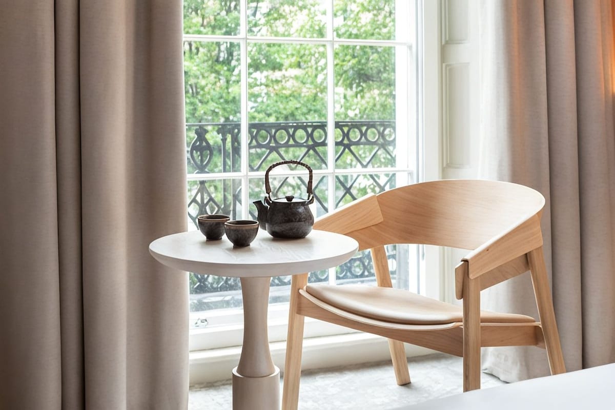 A wooden chair and a small round table with a teapot and cups on it, positioned by a window with a view of greenery.