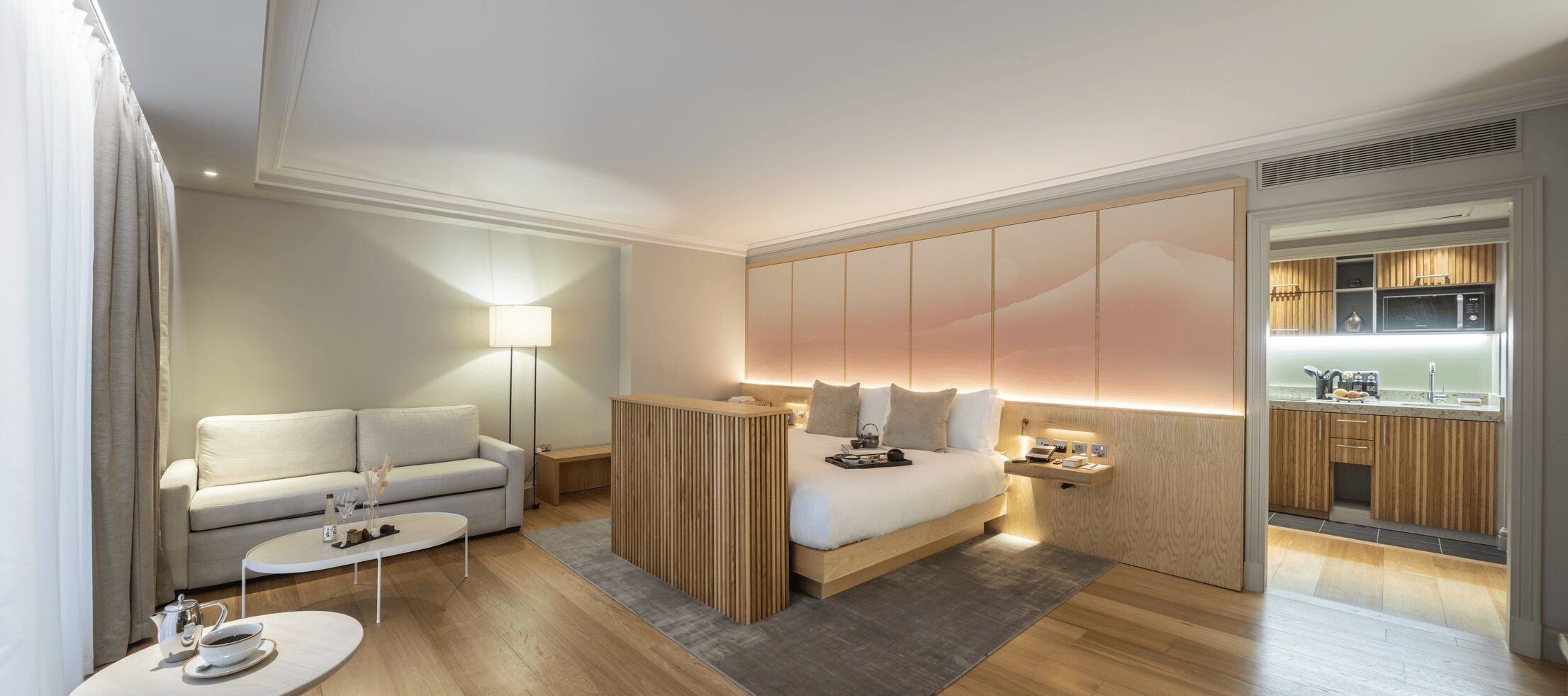 Ein modernes Hotelzimmer mit einer Couch, einem Bett, einem Nachttisch, einer Lampe und Blick zur Küche.