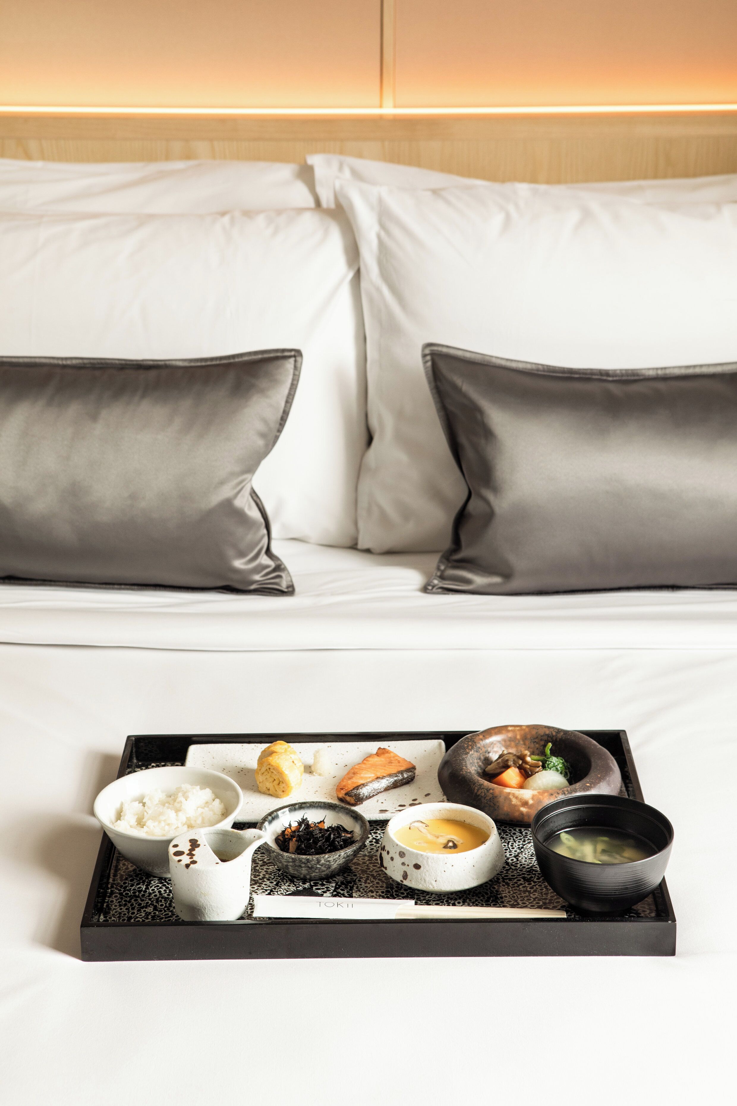 A neatly arranged breakfast tray on a bed with white linens and grey pillows.
