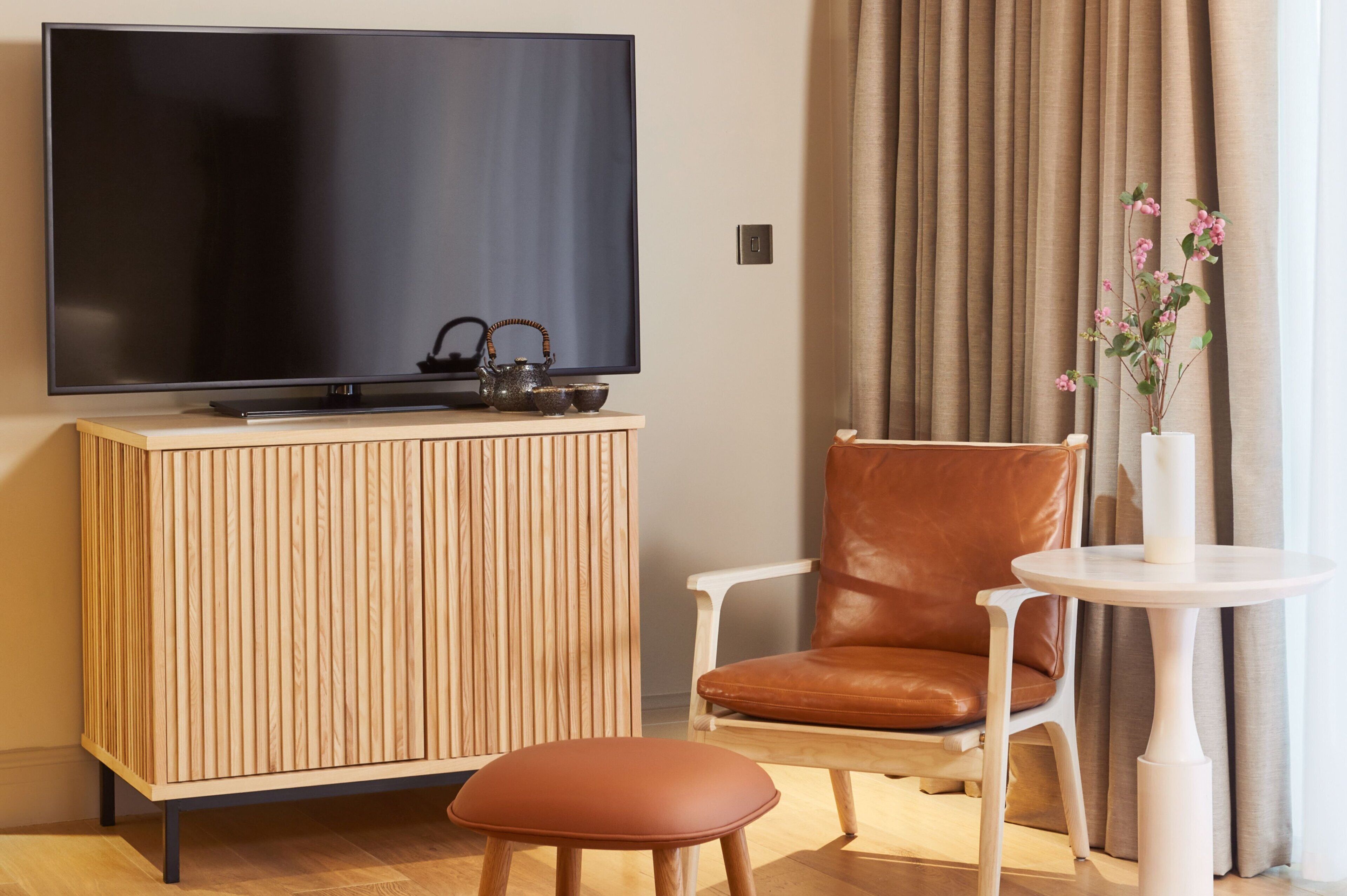 Ein modernes Zimmer mit Flachbildfernseher, einem Holzkabinett, einem braunen Ledersessel, einem kleinen runden Tisch und einer Vase mit Blumen.