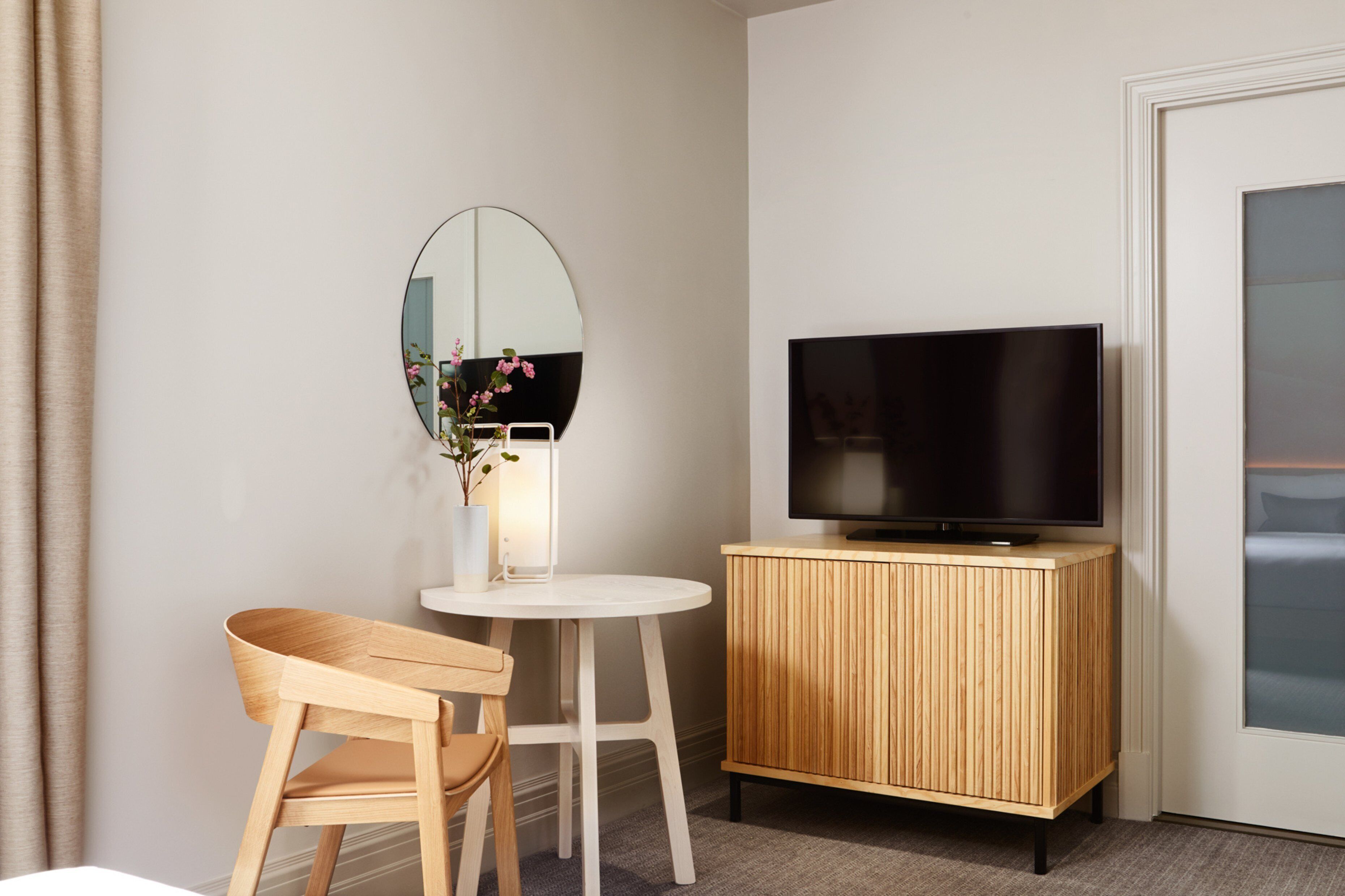 A hotel room with a wooden chair, a small round table, a television on a wooden cabinet, and a mirror on the wall.