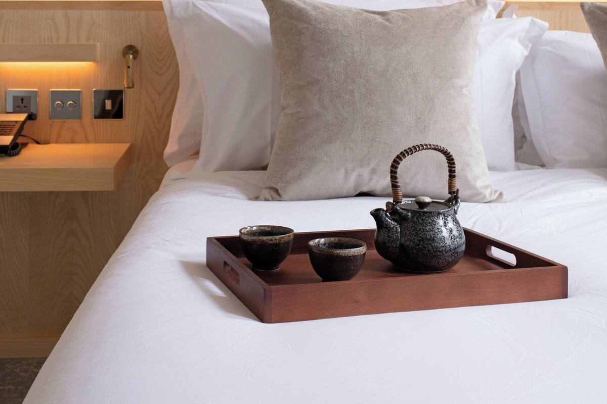 A neatly made bed with a tray of tea and a teapot, a bedside table with a lamp, and a wall-mounted light.