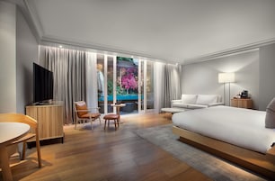 Ett modernt hotellrum med en stor säng, en TV, en sittgrupp med bord och stolar samt utsikt över en trädgård genom fönstret.