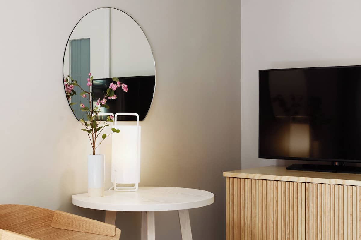 A modern room with a round table, a wooden chair, a television on a wooden cabinet, a mirror on the wall, and a vase with flowers.