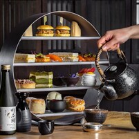 A tea service with a teapot pouring into a cup, surrounded by various pastries and desserts on a tiered stand.