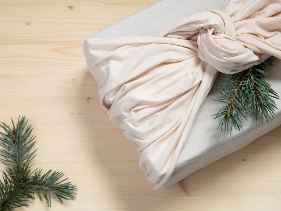 A wrapped gift with a beige cloth tied in a bow, placed on a wooden surface with pine branches.
