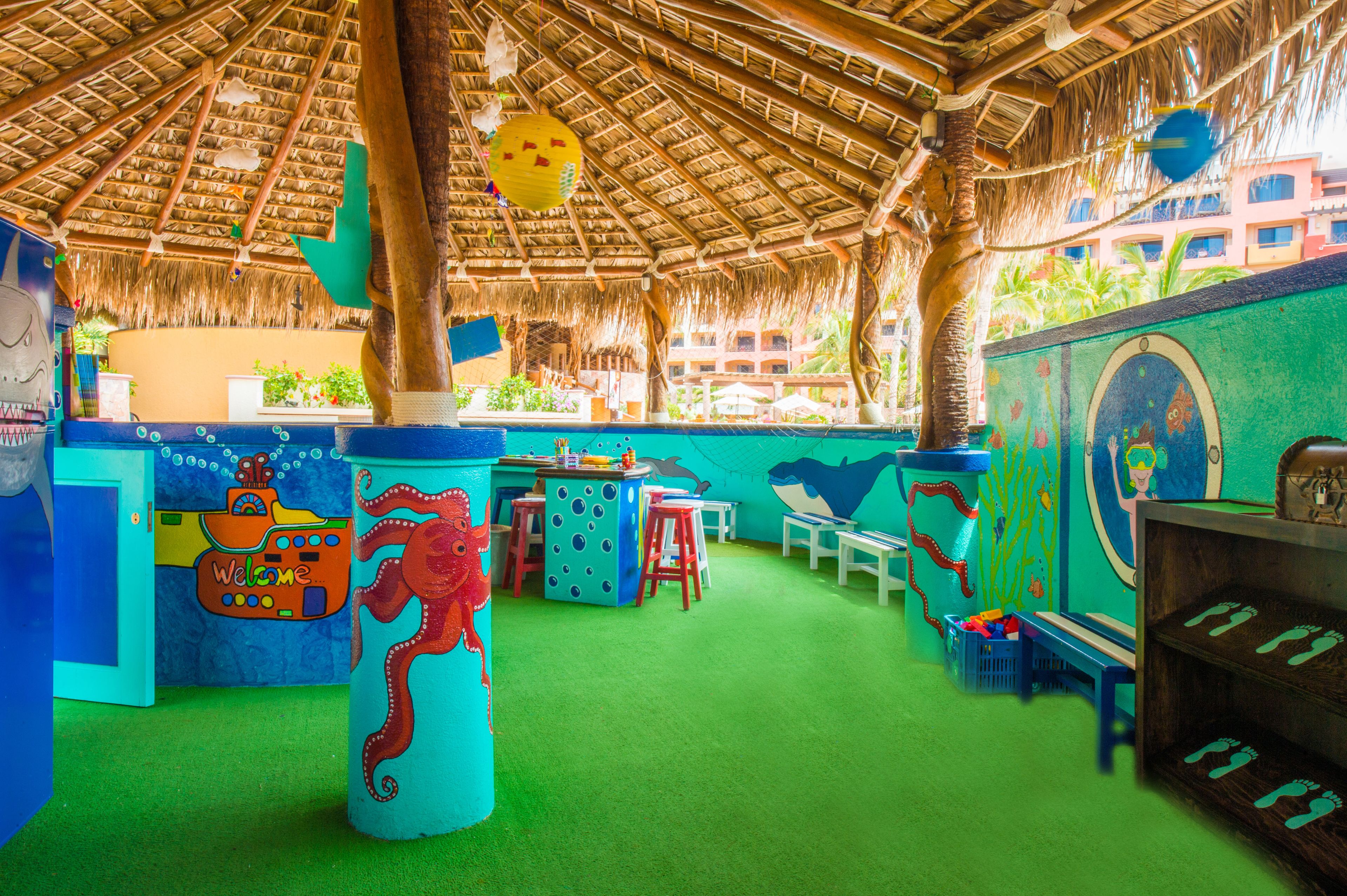 A colorful play area with a thatched roof, green flooring, and various play structures including a large octopus figure and a submarine.