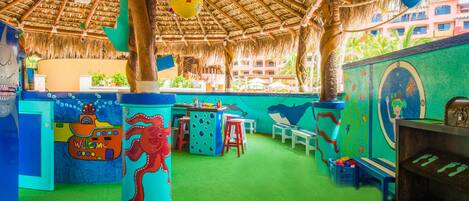 A colorful play area with a thatched roof, green flooring, and various play structures including a large octopus figure and a submarine.