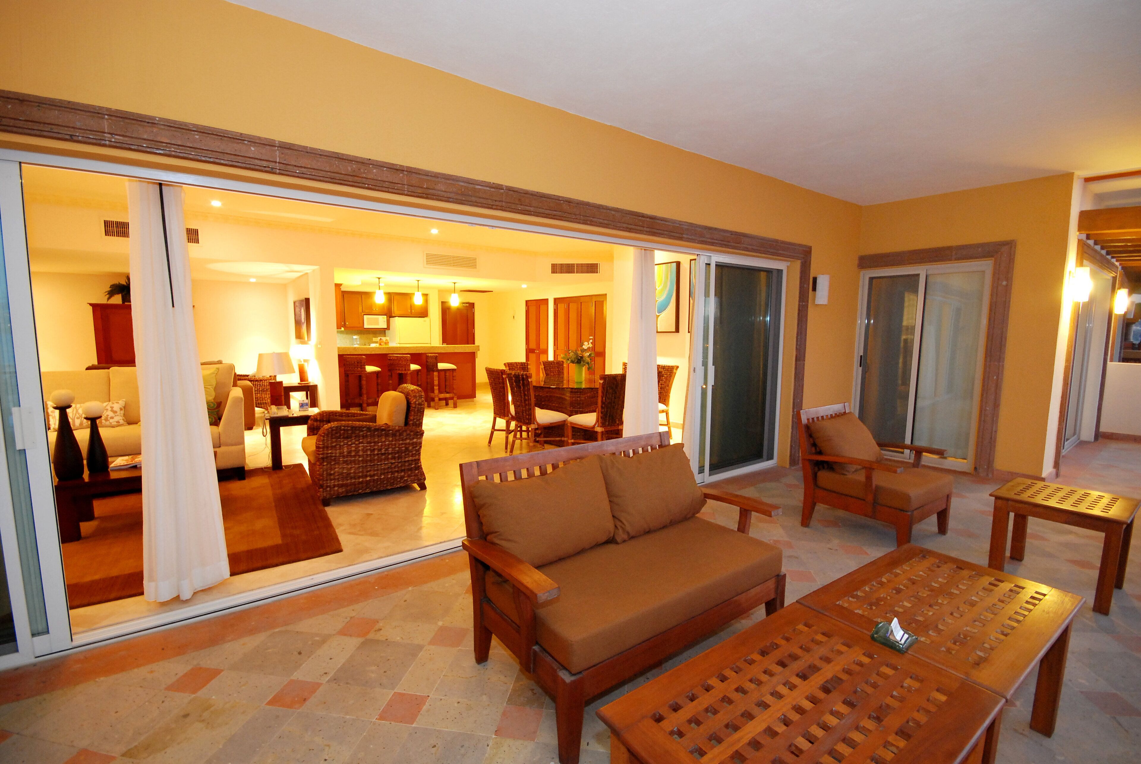A spacious living area with a sofa, chairs, and a wooden coffee table. A sliding glass door leads to a kitchen and dining area.