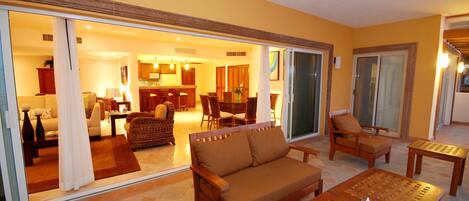 A spacious living area with a sofa, chairs, and a wooden coffee table. A sliding glass door leads to a kitchen and dining area.