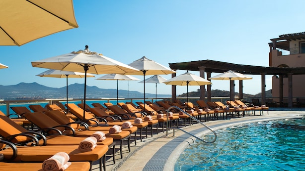 A poolside area with sun loungers and umbrellas, a clear blue pool, and a view of distant mountains.